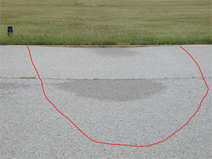 Photo showing a large-severity depression on the edge of an asphalt pavement.            A stringline has been stretched across the depression to illustrate the depth, and some water staining on the pavement surface is noticeable at the center of the depression.            A red line has also been drawn on the photo to outline the area with the depression.